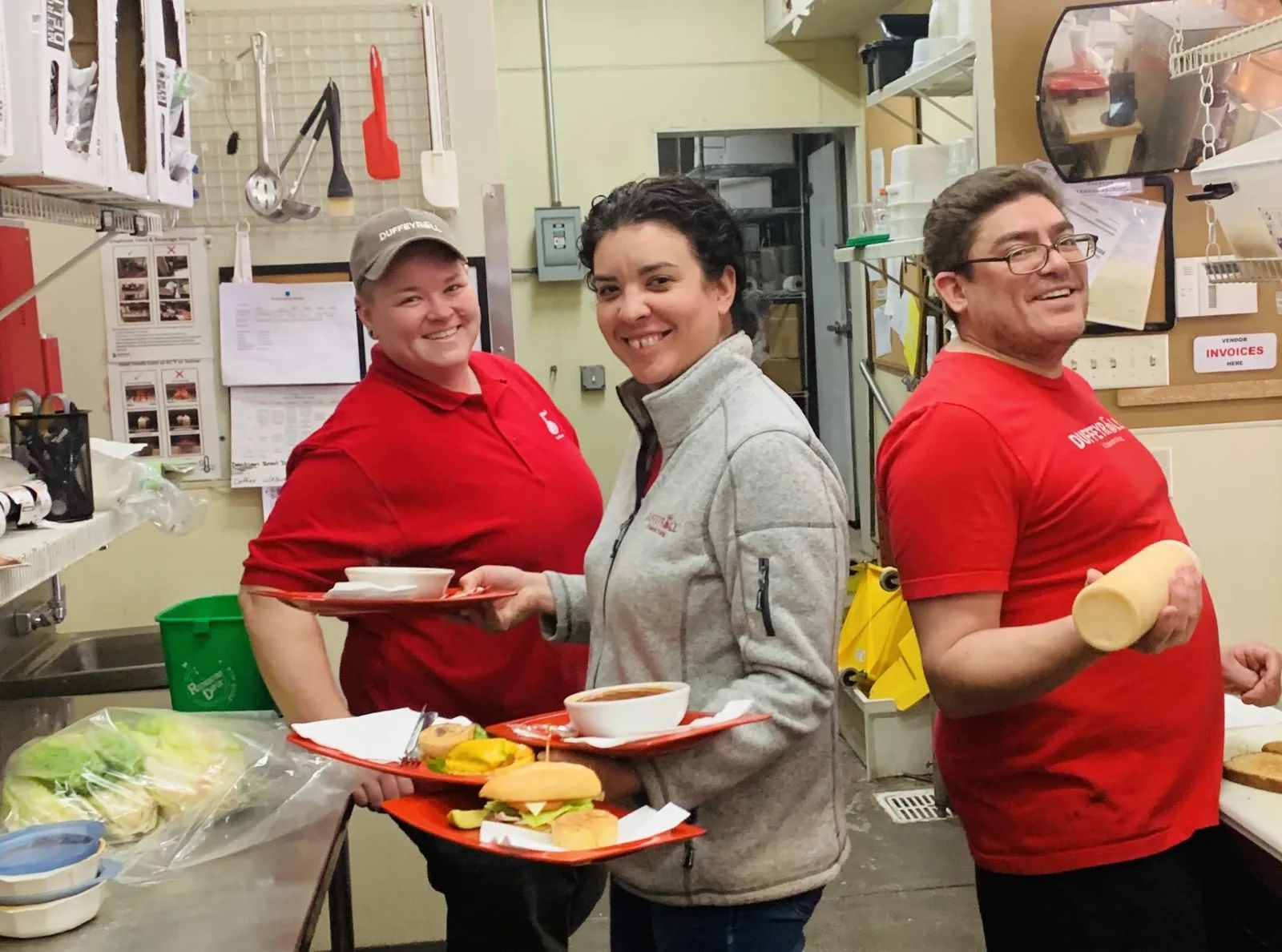 Three Duffeyrolls team members smiling in the café kitchen, two of them carrying trays of food.